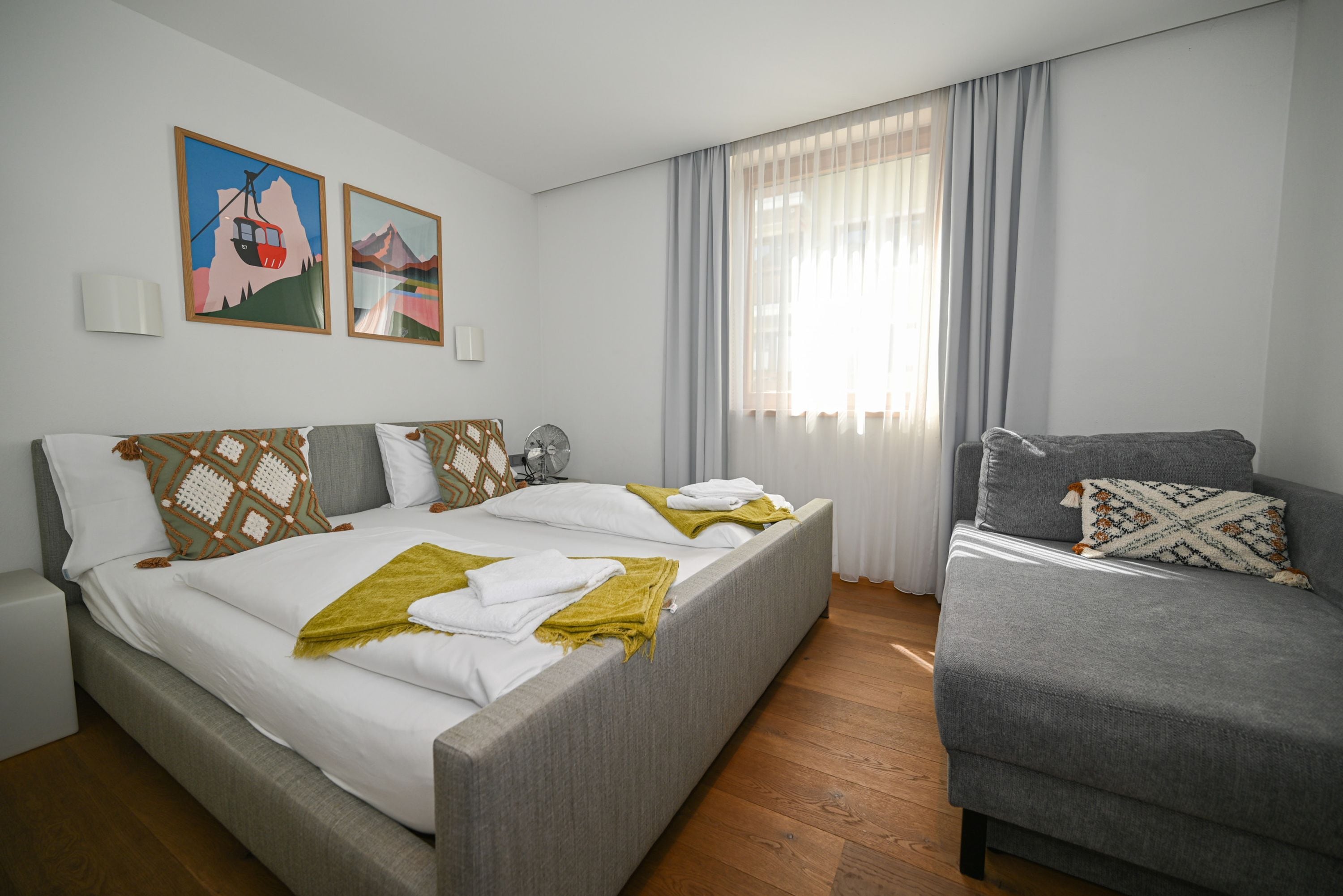 Une chambre d’hôtel avec un lit, un canapé, un ventilateur et deux tableaux accrochés au mur.