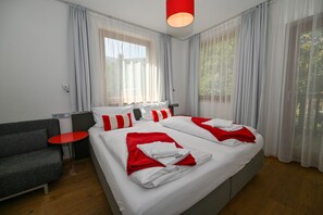 Une chambre d’hôtel moderne avec un grand lit, un canapé gris, une petite table rouge et une fenêtre ornée de rideaux légers.