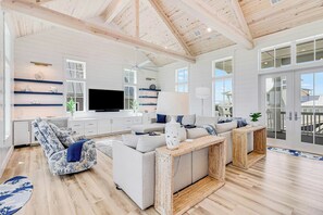 A spacious living room with a high ceiling, built-in shelving, a flat-screen TV, and a large window. - Cattail Castle | Water Views | Steps to Beach (Port Aransas)