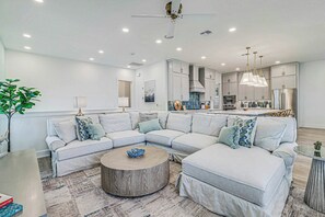 A modern living room with a sectional sofa, a round coffee table, and a kitchen area with grey cabinets and stainless steel appliances. - 'Merica Estate | Steps to Beach | Game Room (Port Aransas)