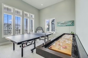 A room with a ping pong table, foosball table, and a large window with a view of houses. - A Wave From It All | Gulf Views | Steps to Beach (Port Aransas)