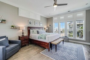 Ein Schlafzimmer mit einem großen Bett, Nachttischen, einer Bank, einem Deckenventilator und Blick durch große Fenster.