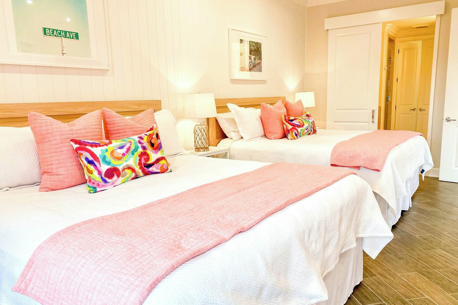 A hotel room with two beds, each with pink bedding and colorful pillows, a wooden headboard, a framed picture on the wall, and a door with a light switch.