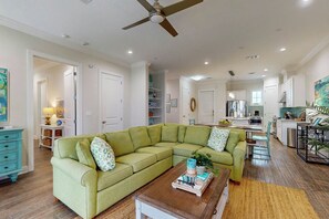 A spacious living room with a green sectional sofa, a wooden coffee table, and a dining area with a kitchen in the background. - Birdie Beach | Steps to Beach | Resort Amenities (Port Aransas)