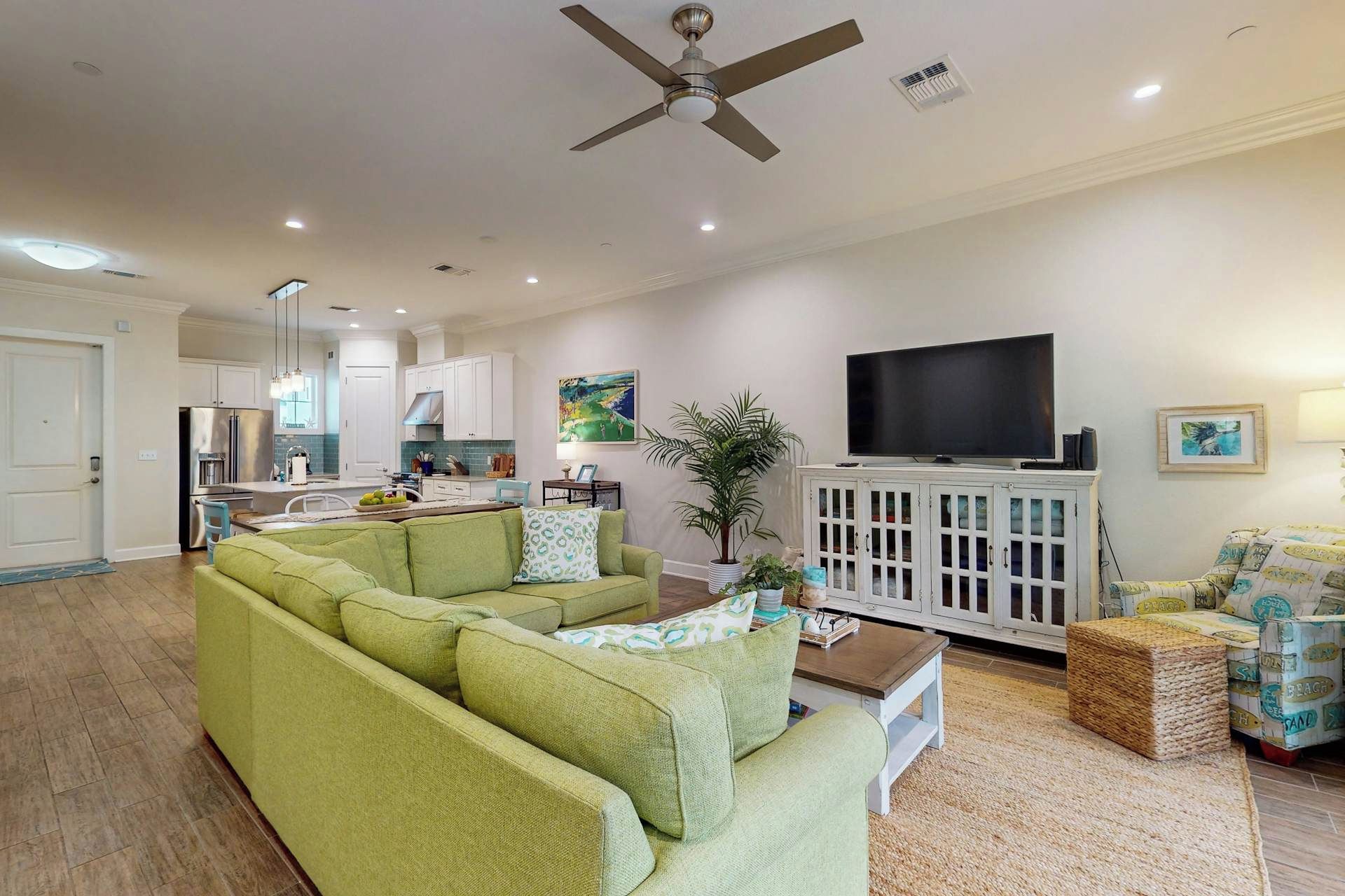 A living room with a green sofa, a television on a cabinet, and a kitchen in the background.