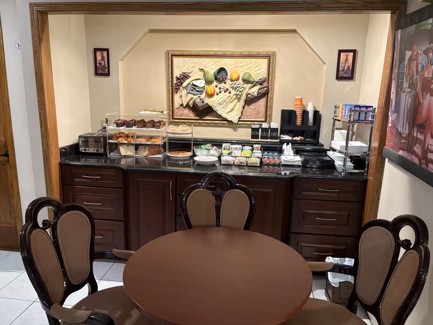 A breakfast bar with various food items, a framed artwork above it, and a dining area with chairs and a table.