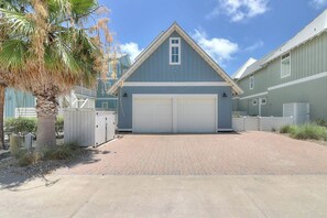 A two-story building with a garage entrance, a brick driveway, and a palm tree. - Life's Good | Gulf Views | Steps to Beach (Port Aransas)