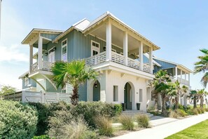 A two-story house with a balcony, a covered porch, and a well-maintained garden with palm trees and shrubs. - Kokomo J's | Gulf Views | Steps to Beach (Port Aransas)