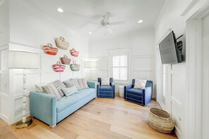 Un salón amplio con un sofá azul claro, dos sillones azules, un ventilador de techo y un televisor montado en la pared.