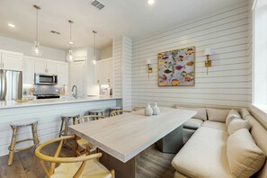 A modern kitchen with white cabinetry, a central island with bar stools, and a dining area with a light-colored table and beige sofa. - Casita Bonita | Gulf Views | Pet Friendly (Port Aransas)