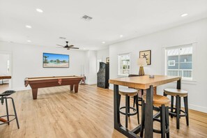 A spacious room with a pool table, bar stools, and a ceiling fan. - Adventures At Sunrise | Gulf Front | Game Room (Port Aransas)
