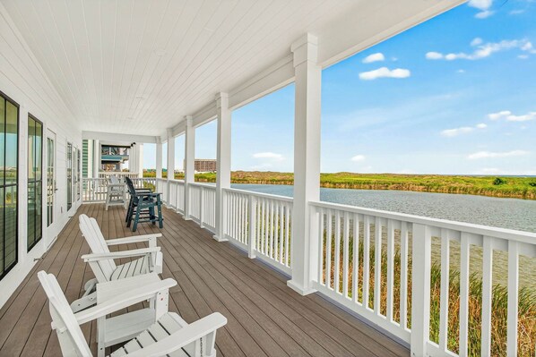 A covered porch with white railings, a table with chairs, and a view of a water body and landscape. - Adventures At Sunrise | Gulf Front | Game Room (Port Aransas)