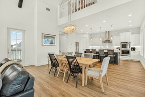 A spacious dining area with a high ceiling, a central chandelier, and a view of the kitchen in the background. - Adventures At Sunrise | Gulf Front | Game Room (Port Aransas)