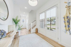 A hallway with a wooden floor, white walls, a staircase, and a large window with a view of palm trees and buildings. - Ocean Haven | Water Views | Private Elevator (Port Aransas)