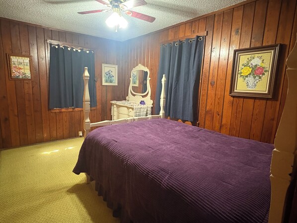 A bedroom with wooden paneling, a bed with a purple bedspread, a dresser, a mirror, and two windows with curtains.