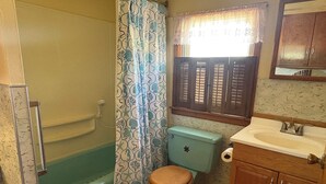Une salle de bain avec une baignoire, des toilettes, un lavabo et une fenêtre avec des volets.