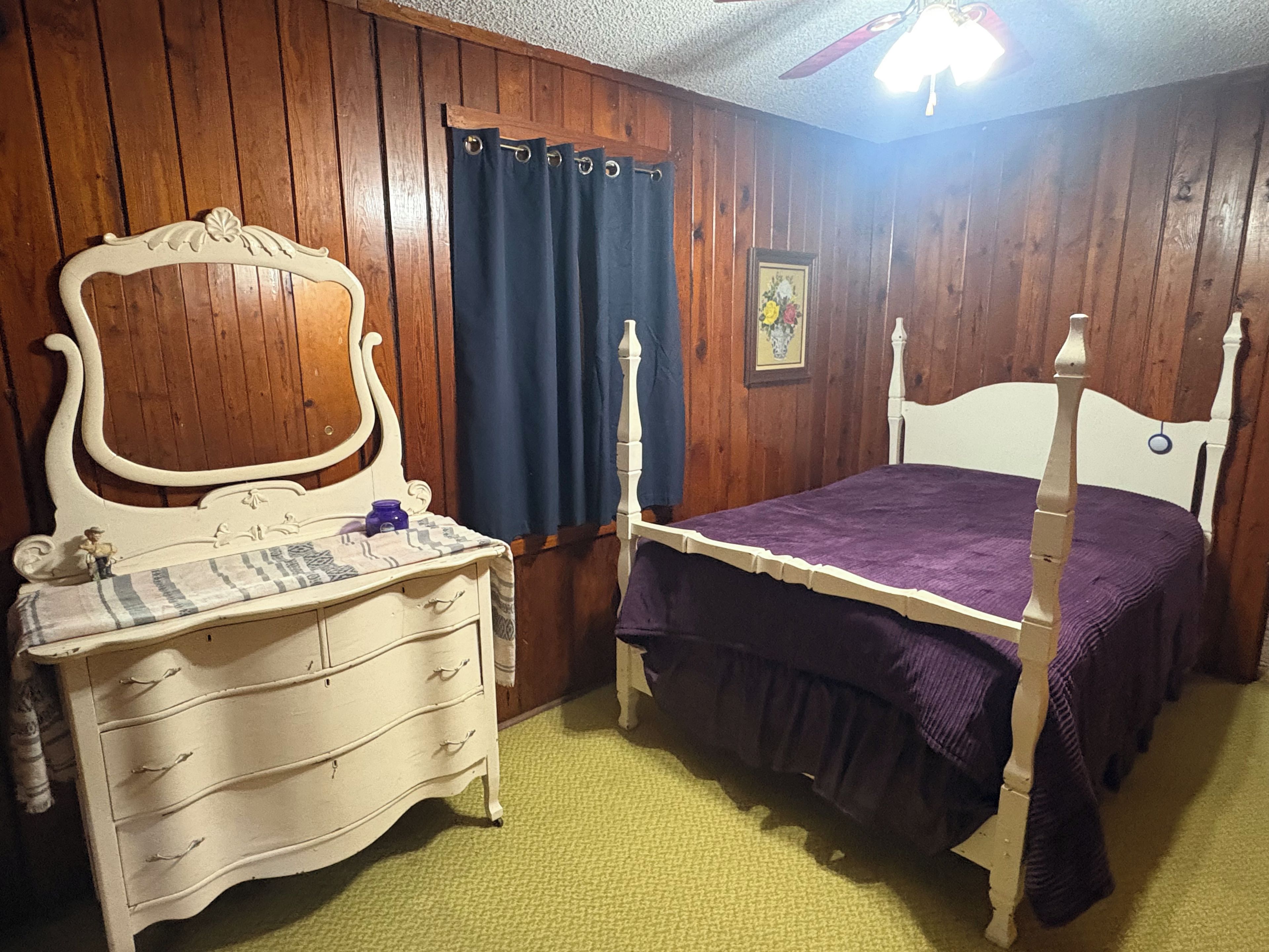 A bedroom with a wooden paneled wall, a bed with a white headboard, a dresser with drawers, a nightstand, and a blue curtain.