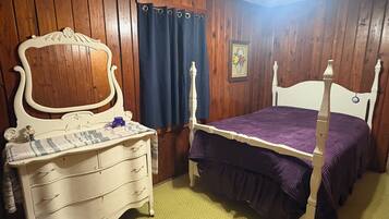 A bedroom with a wooden paneled wall, a bed with a white headboard, a dresser with drawers, a nightstand, and a blue curtain.