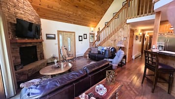 A cozy living room with a stone fireplace, a leather sofa, a wooden dining table, and a staircase leading to an upper level.