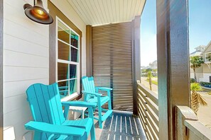 A covered porch with two turquoise Adirondack chairs and a table, a window with blinds, and a view of a residential street. - Coastal Vista Retreat | Pet Friendly (Port Aransas)