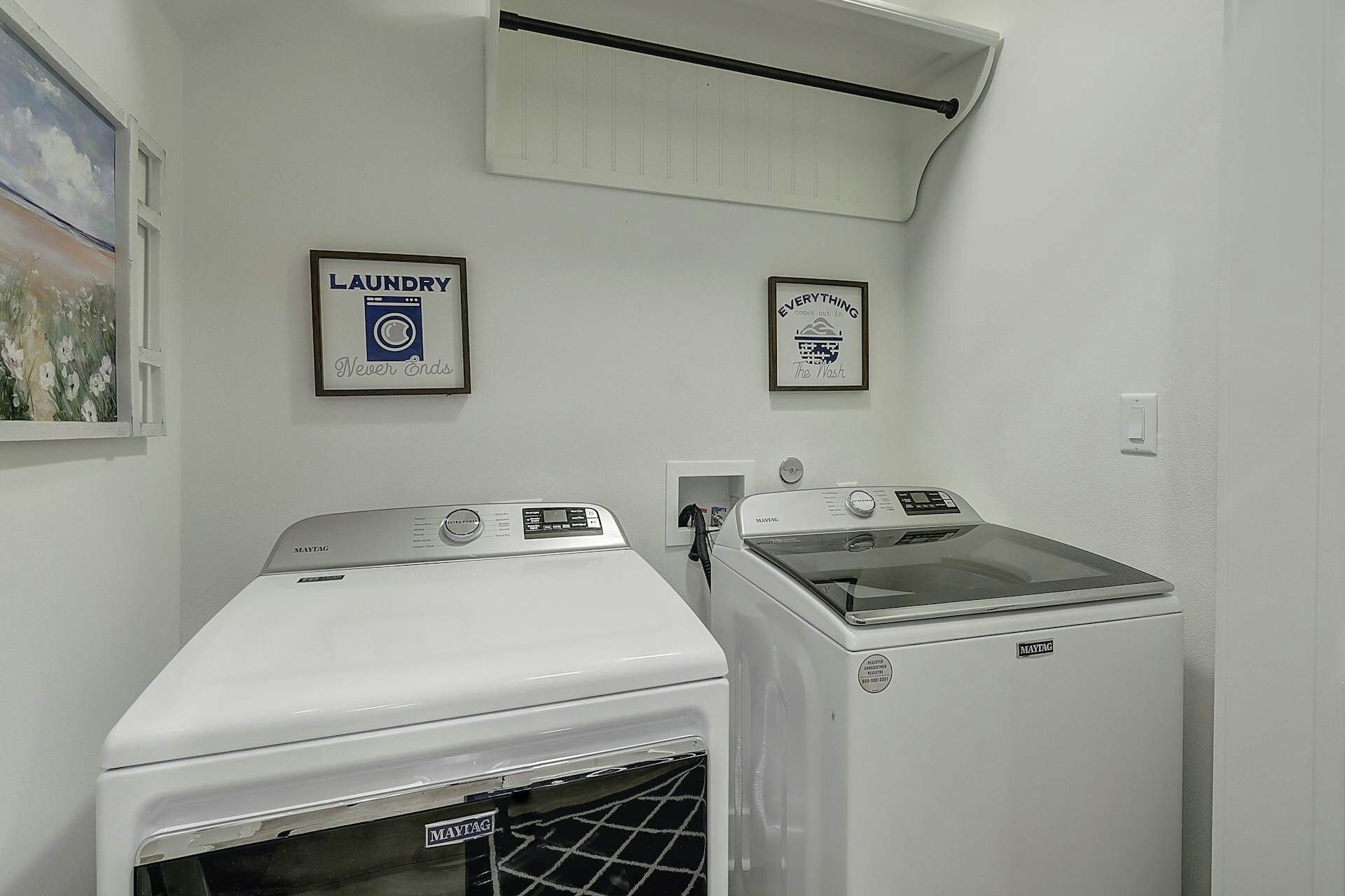 Un lavadero con dos lavadoras y secadoras Maytag, un cuadro de flores y dos carteles enmarcados en la pared.