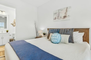 A bedroom with a bed, a nightstand, a lamp, a wall-mounted painting, and a bathroom visible in the background. - Driftwood Cottage | Golf Views | Pet Friendly (Port Aransas)