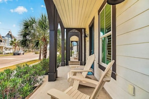 A covered porch with Adirondack chairs, a grill, and a view of a street with houses. - Driftwood Cottage | Golf Views | Pet Friendly (Port Aransas)