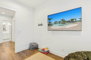 A room with a large framed picture of a pool area, a bean bag chair, and a rug on the floor. - Driftwood Cottage | Golf Views | Pet Friendly (Port Aransas)