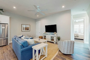 A living room with a sofa, a television, a ceiling fan, and a kitchen area with a refrigerator. - Beach Please | Steps to Beach | Resort Amenities (Port Aransas)