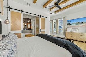 A bedroom with a bed, a sliding door leading to a bathroom, a ceiling fan, and a window with curtains. - Gone Breeze | Pet Friendly | Steps to Beach (Port Aransas)