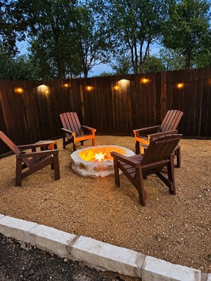 A fire pit surrounded by four Adirondack chairs in a gravel area, with a wooden fence and string lights in the background.