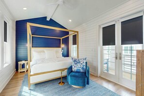 A bedroom with a four-poster bed, a round gold side table, a blue upholstered chair, and a large window with a view. - A Shore Thing | Gulf Views | Steps to Beach (Port Aransas)