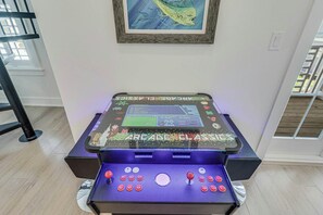 Una máquina de videojuegos retro con pantalla y varios botones, ubicada sobre una mesa en una habitación con ventana y un cuadro en la pared.