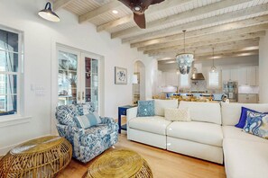 A spacious living room with a white sofa, blue patterned armchair, and wicker ottomans. A kitchen with modern appliances and a dining area with blue and white checkered tablecloths is visible in the background. - A Shore Thing | Gulf Views | Steps to Beach (Port Aransas)