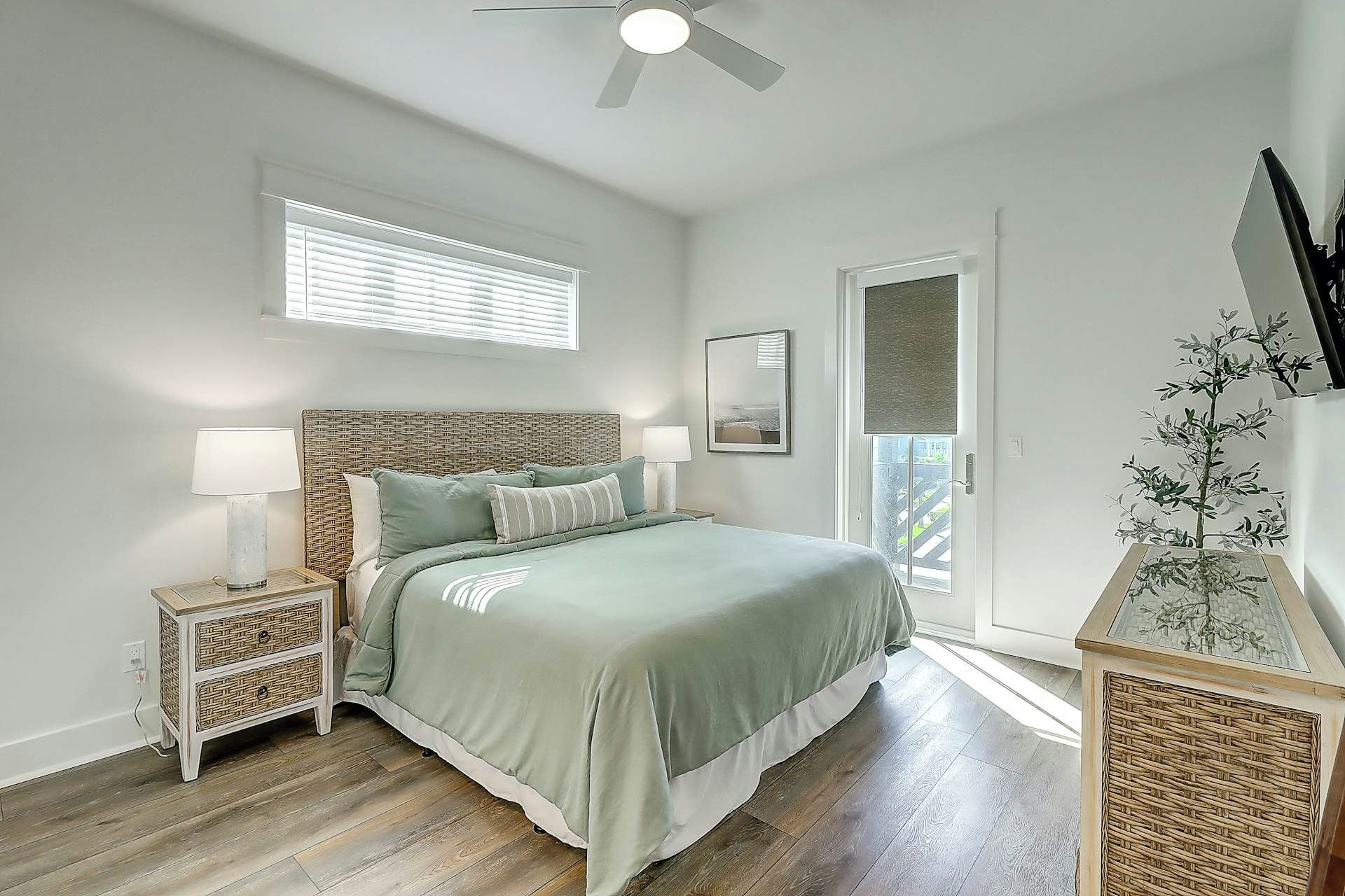 Une chambre à coucher comprenant un lit, des tables de chevet, un ventilateur de plafond, une fenêtre avec des stores, un tableau encadré et une plante en pot.