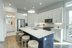 A modern kitchen with white cabinetry, stainless steel appliances, a large island with stools, and pendant lighting. - Beaches And Bunkers | Golf Views | Pet Friendly (Port Aransas)