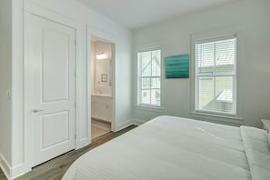 A bedroom with a bed, a door, a bathroom, and two windows with blinds. - Beaches And Bunkers | Golf Views | Pet Friendly (Port Aransas)