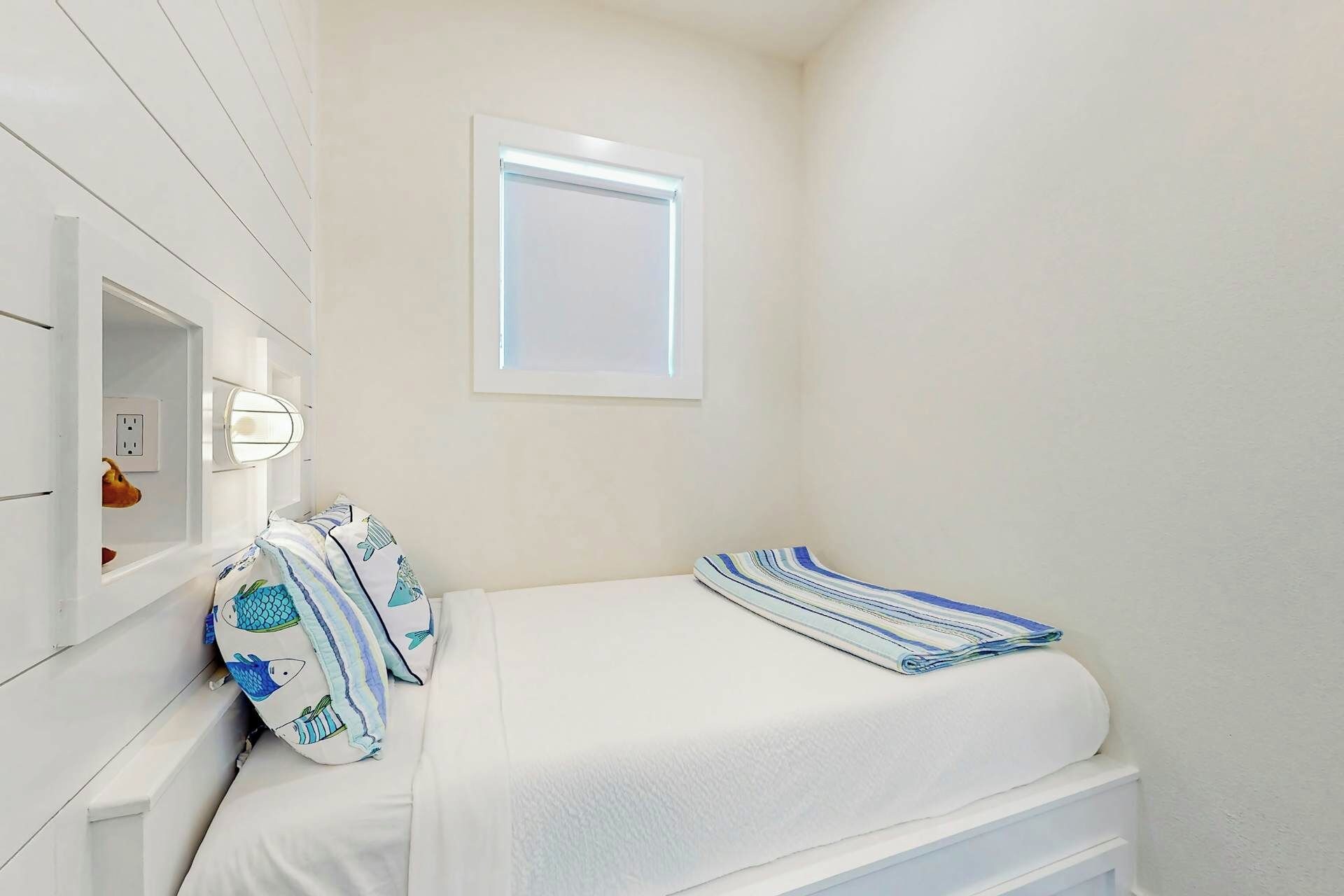 Ein kleines, ordentlich eingerichtetes Schlafzimmer mit einem Einzelbett, einem Nachttisch, einer Wandleuchte und einem Fenster.