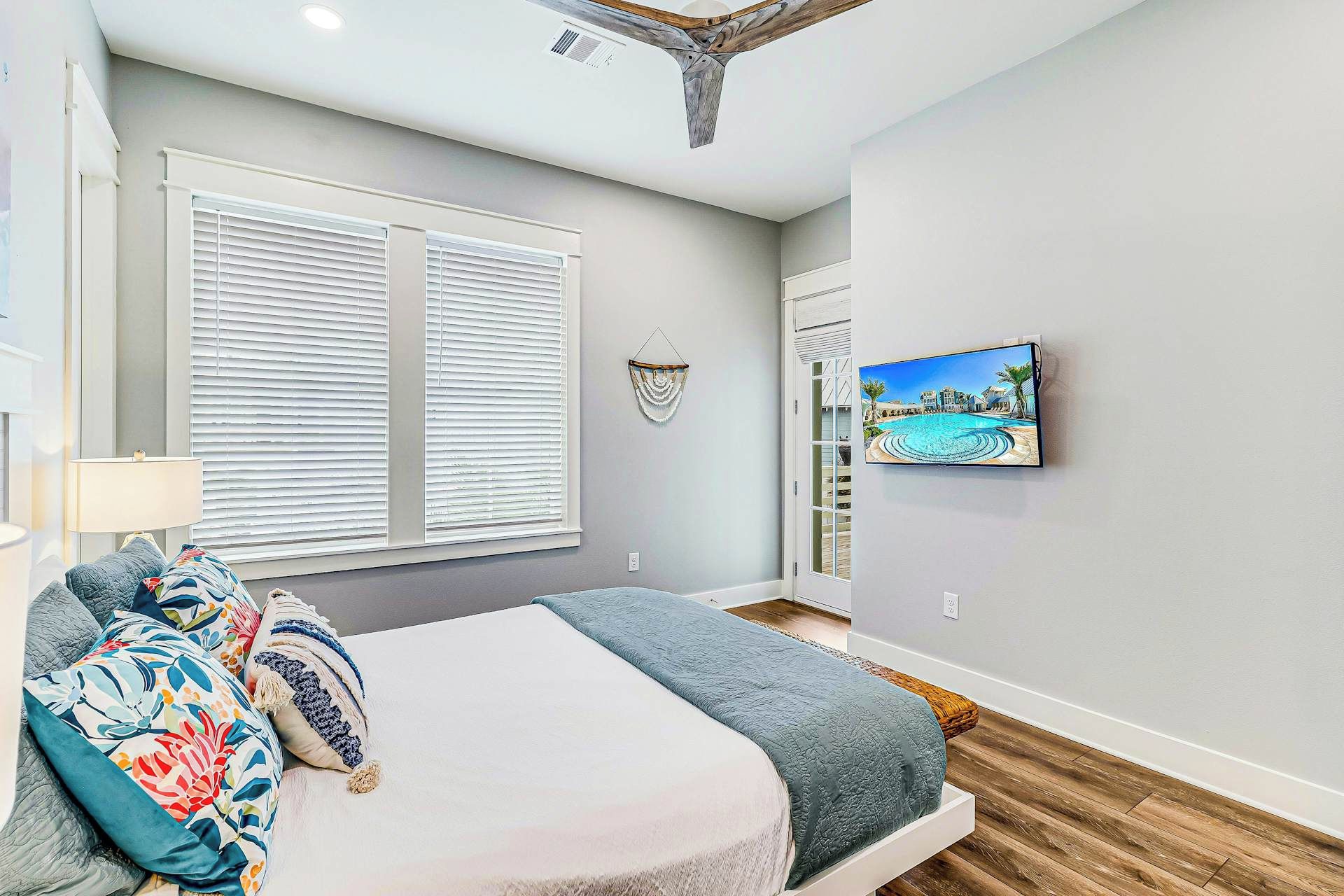 Un dormitorio con cama, ventana con persianas, televisor montado en la pared y puerta que da a un balcón.