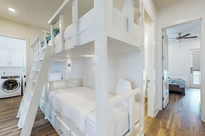 Ein Schlafraum mit Etagenbetten, im Hintergrund eine Waschmaschine, im angrenzenden Raum ein Deckenventilator und eine Tür zu einem weiteren Zimmer.