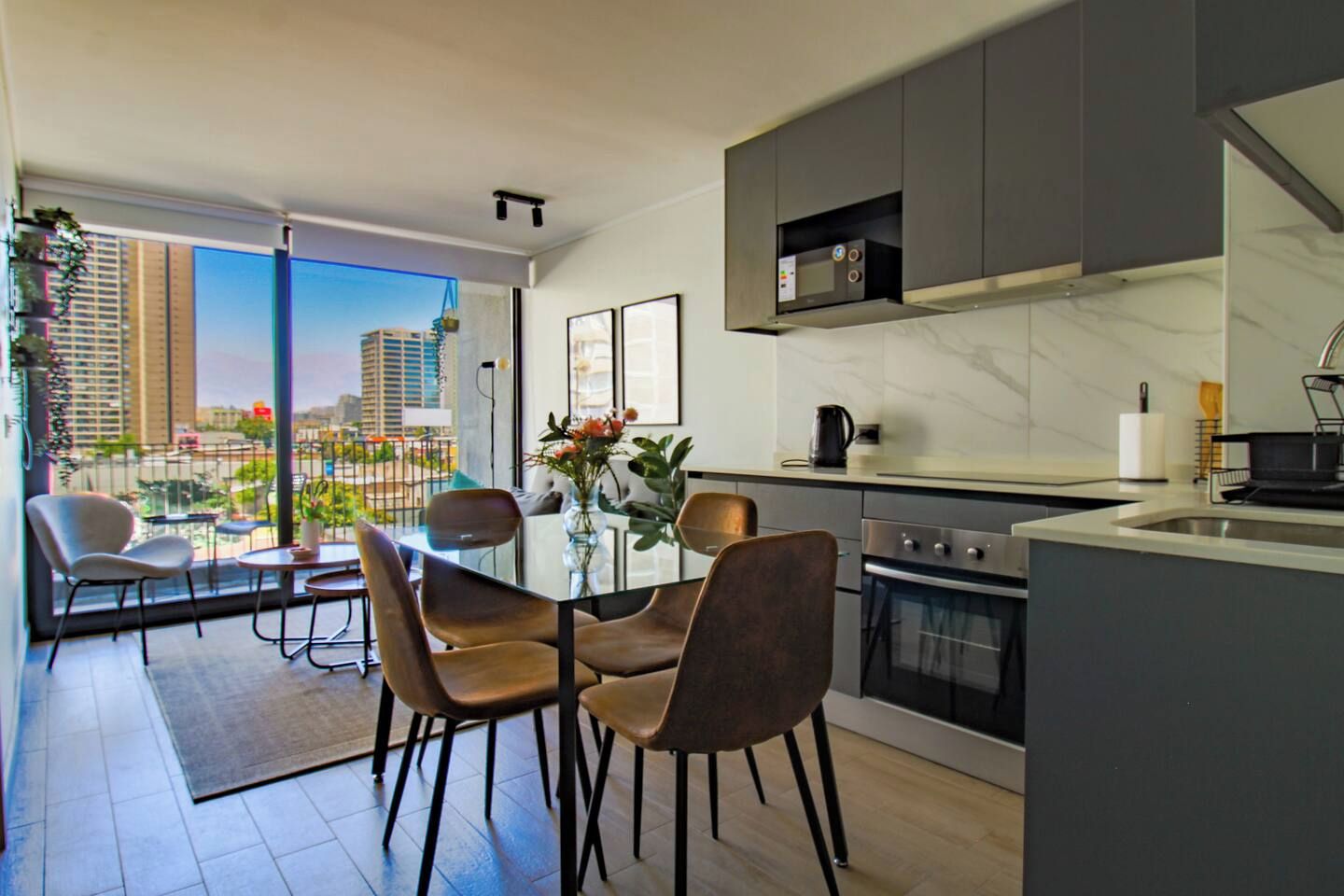 A modern kitchen with a built-in oven, a dining area with a glass table and chairs, and a view of the city through large windows.