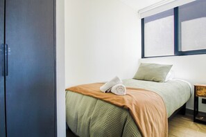 Ein Einzelbett mit einer braun-grünen Tagesdecke, ein aufgerolltes weißes Handtuch und ein grünes Kopfkissen in einem Zimmer mit weißen Wänden und einem Fenster.