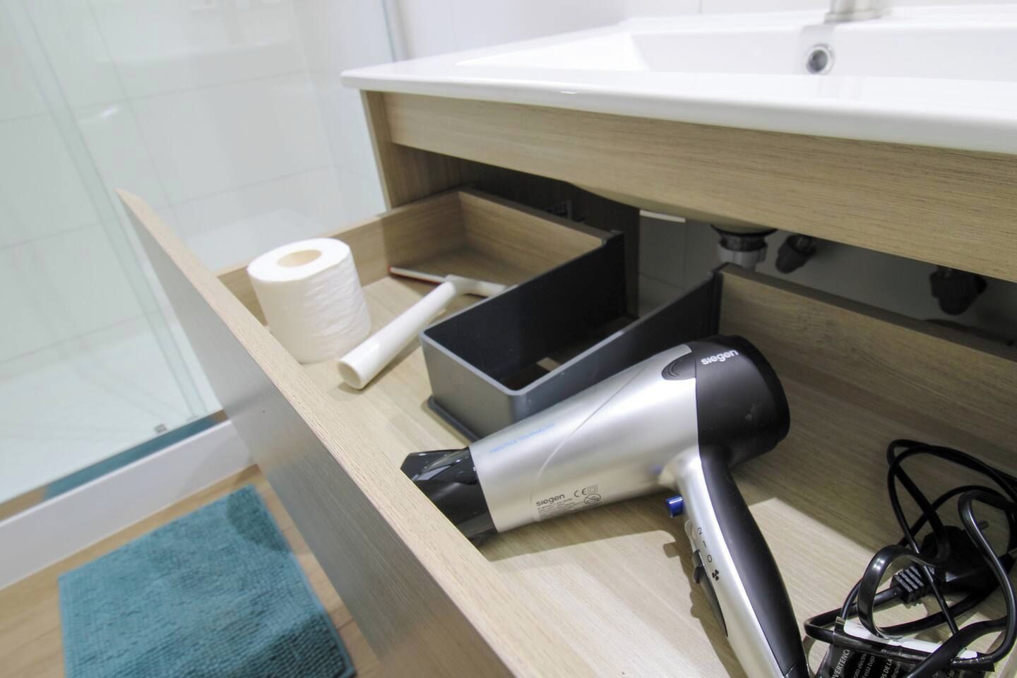 Kylpyhuone, jossa on puinen pesuallas, hiustenkuivaaja, wc-paperirulla ja musta latauskaapeli.