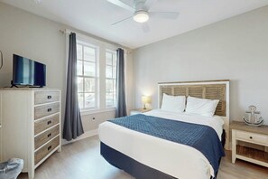A hotel room with a bed, a TV on a dresser, a nightstand with a lamp, and a ceiling fan. - Crab Manor | Steps to Beach | Resort Amenities (Port Aransas)