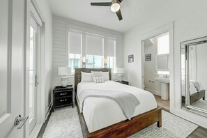 A bedroom with a bed, bedside tables, a ceiling fan, a window, and a bathroom with a sink and mirror. - At Ease | Golf Views | Steps to Beach (Port Aransas)