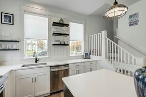 A modern kitchen with white cabinetry, a stainless steel dishwasher, and a large window providing a view of the outside. - At Ease | Golf Views | Steps to Beach (Port Aransas)