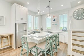 A modern kitchen with white cabinetry, a stainless steel refrigerator, a central island with chairs, and pendant lighting. - Beach Days and Fairways | Golf Views (Port Aransas)