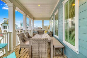 A covered porch with wicker furniture, a wooden table, and a view of neighboring buildings. - Glam Wave | Gulf Views | Steps to Beach (Port Aransas)