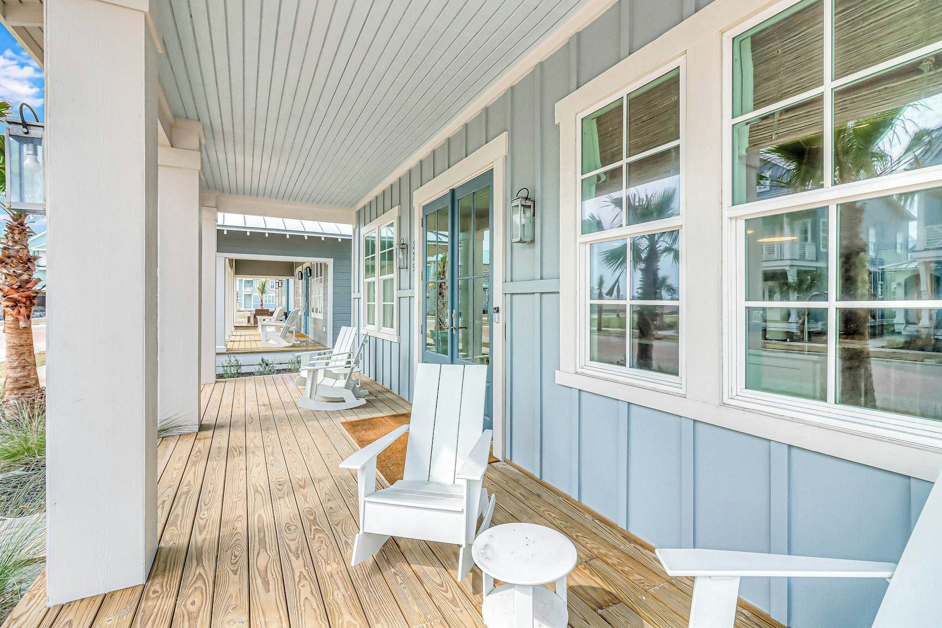 Eine überdachte Veranda mit Holzboden, weißen Adirondack-Stühlen und großen Fenstern.