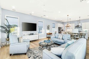 A modern living room with a flat-screen TV, light blue sofas, a coffee table, and a dining area with a chandelier. - Allen's Anchor | Pet Friendly | Steps to Beach (Port Aransas)